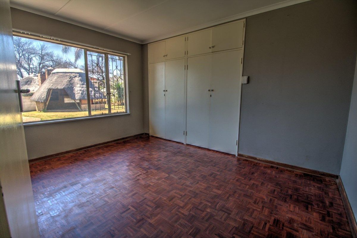 An empty bedroom featuring original parquet flooring, built-in cupboards, and large windows with security bars overlooking the garden.