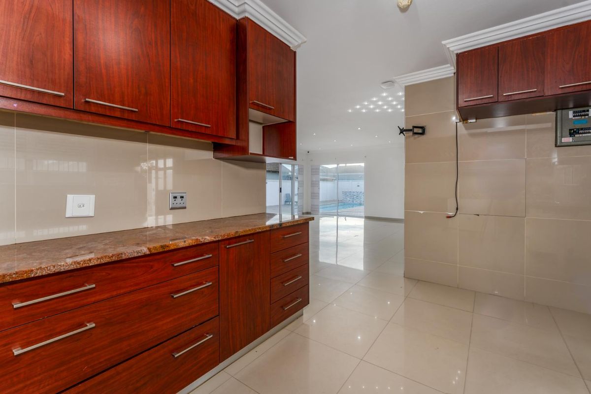A modern kitchen area featuring rich wood-toned cabinetry, granite countertops, and polished floor tiles, leading into an open-plan living space with pool views.