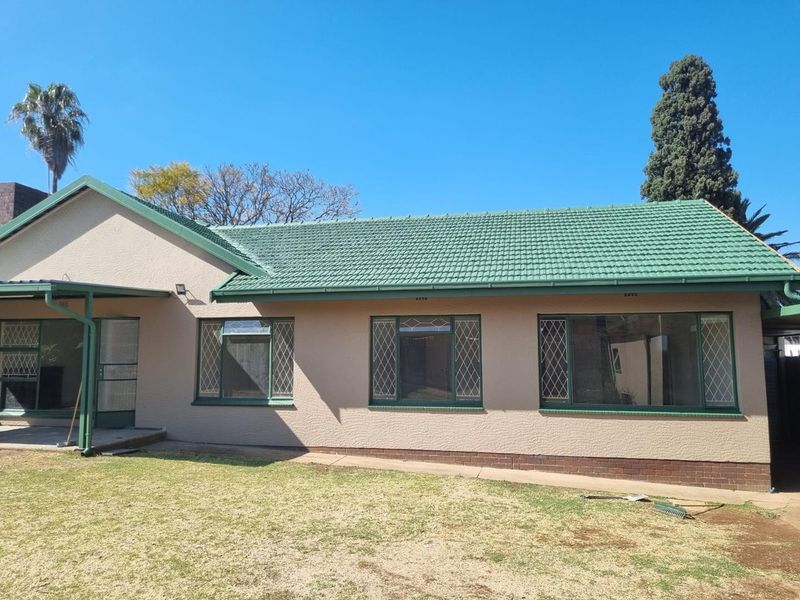 Rear view of a single-story house featuring a green tiled roof, textured beige walls, and a covered patio area overlooking the garden.