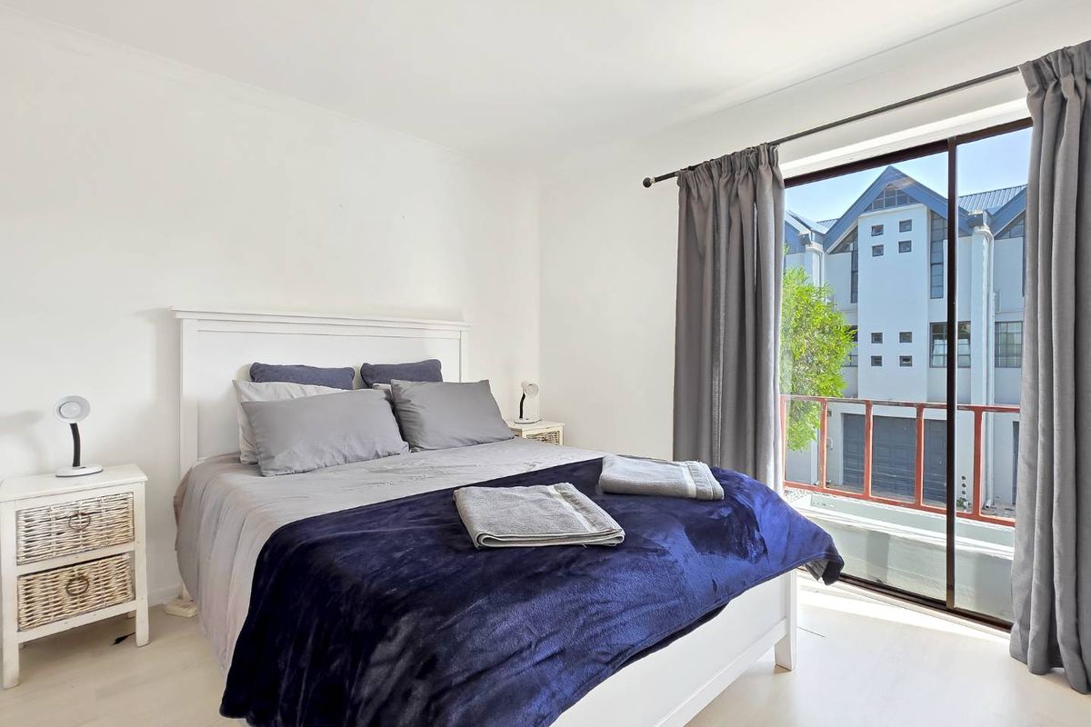 A bright bedroom featuring light-toned flooring and a large sliding glass door that opens to a private balcony with exterior views.