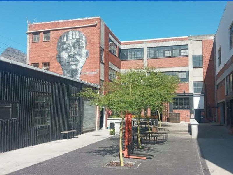 An industrial-style courtyard featuring exposed brick architecture, large steel-framed windows, and a prominent mural, creating a unique mixed-use outdoor space.