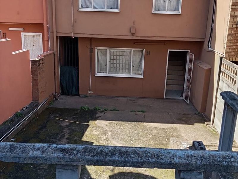 Rear view of the townhouse featuring a concrete patio area, security gates, and a brick boundary wall, showing signs of weathering and maintenance requirements.