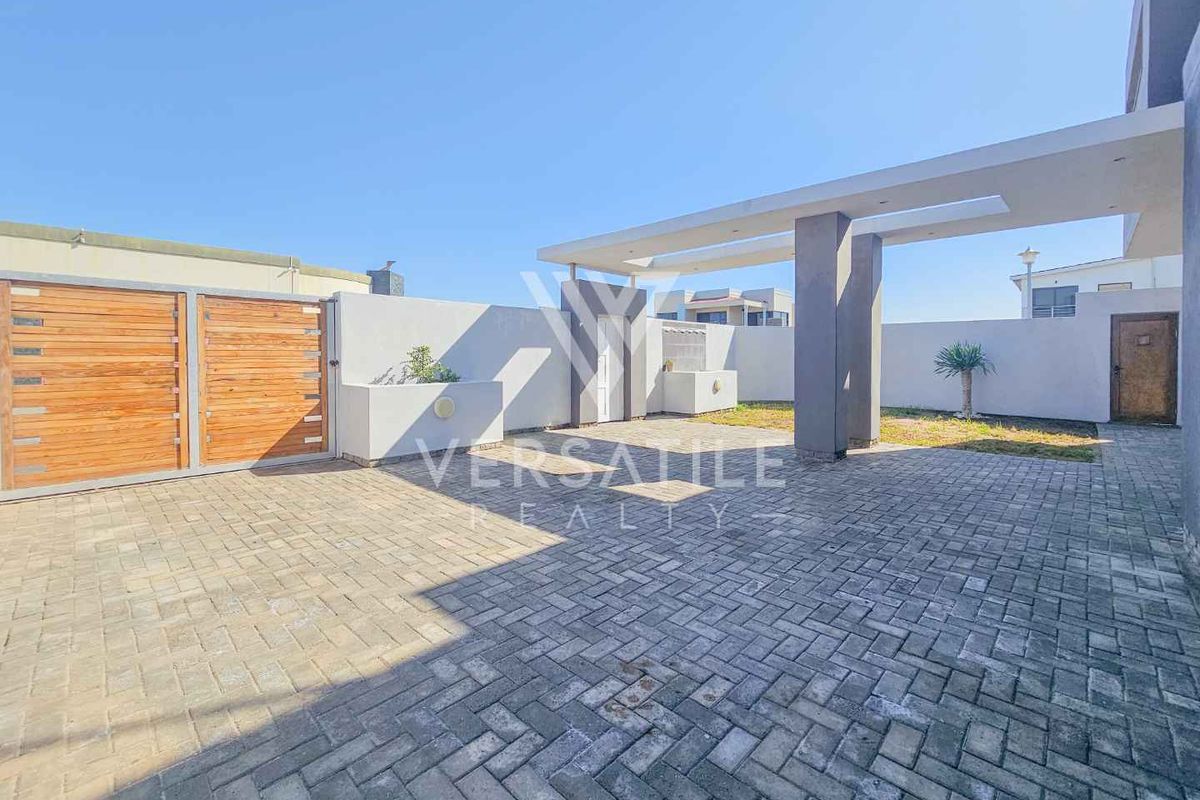 A spacious rear exterior area featuring a paved driveway, a covered patio structure, and secure boundary walls with a timber gate.