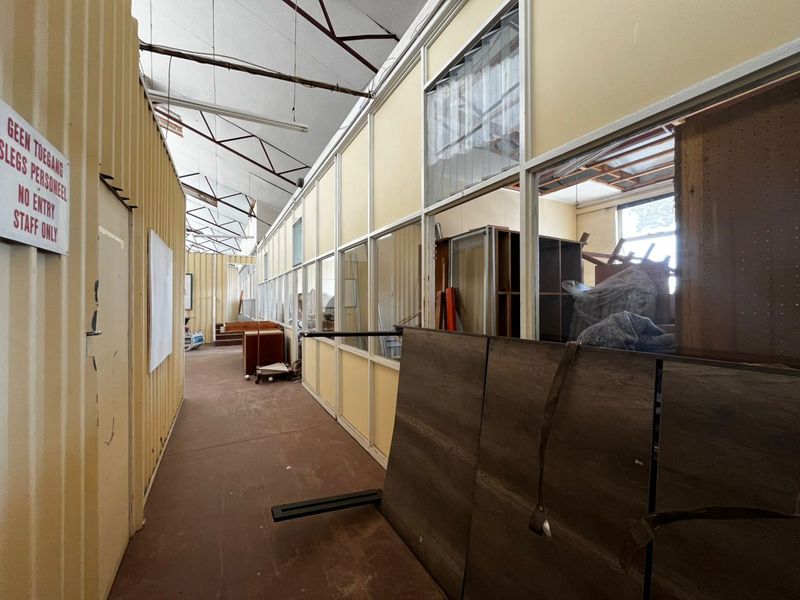 A long industrial-style hallway featuring corrugated metal walls, partitioned office glazing, and exposed roof trusses, currently used for storage.