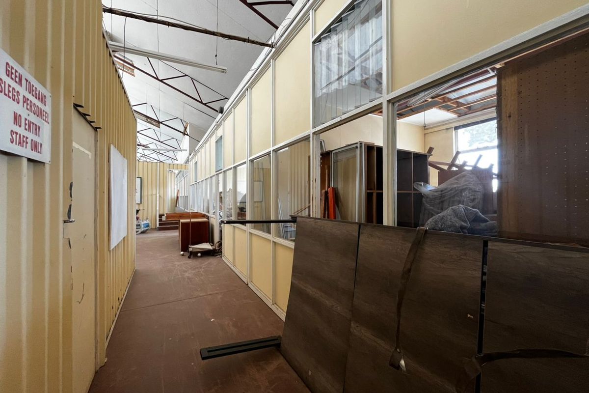 A long industrial-style hallway featuring corrugated metal walls, partitioned office glazing, and exposed roof trusses, currently used for storage.