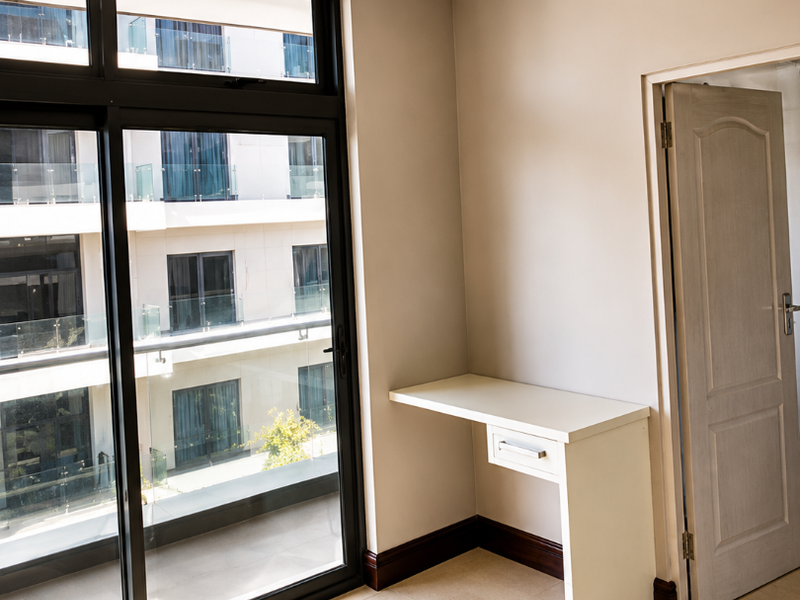 A bright, empty bedroom featuring tiled flooring, crown molding, a built-in desk, and a large sliding glass door providing access to a balcony.