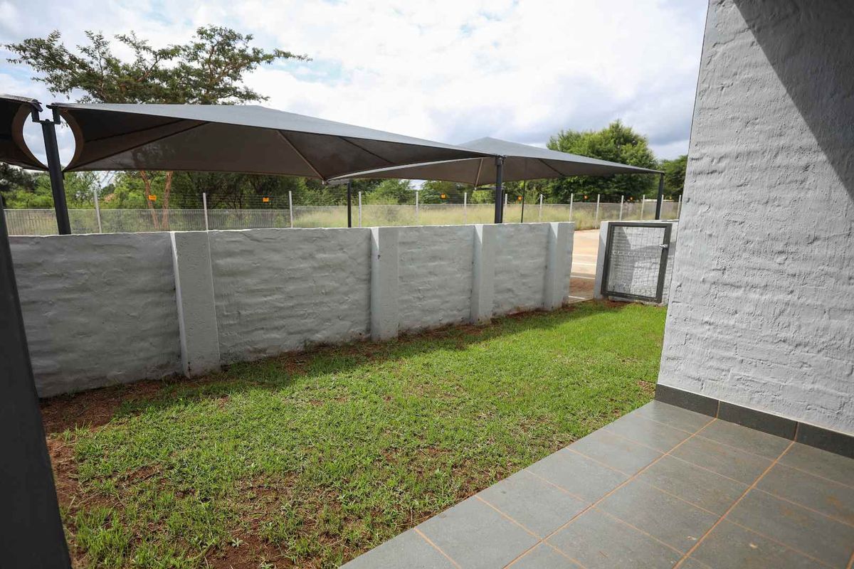 A private rear garden area featuring a tiled patio, a well-maintained lawn, and a shaded parking structure enclosed by a boundary wall.