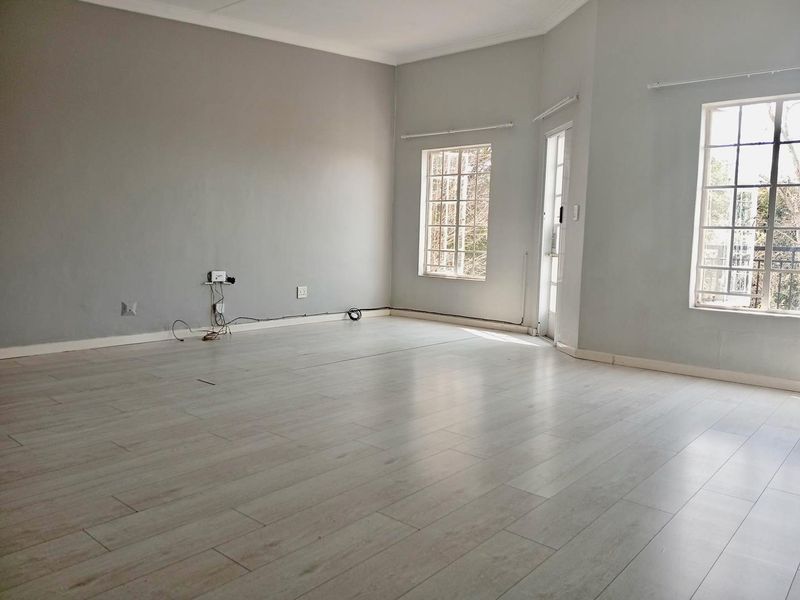 A bright, empty living room featuring light-toned laminate flooring, neutral walls, coving, and multiple windows providing natural light.