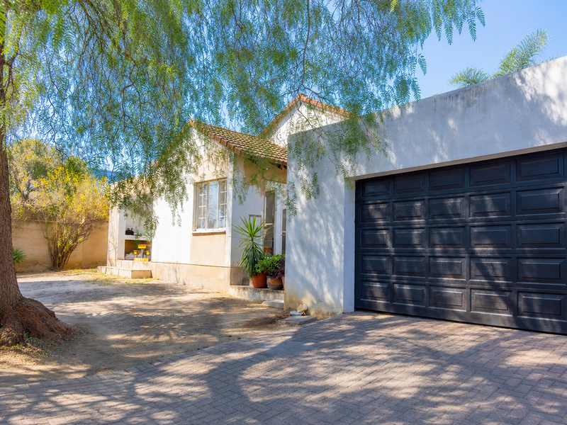 Front elevation of the house featuring a paved driveway, a dark garage door, and a built-in outdoor braai area under a mature tree.