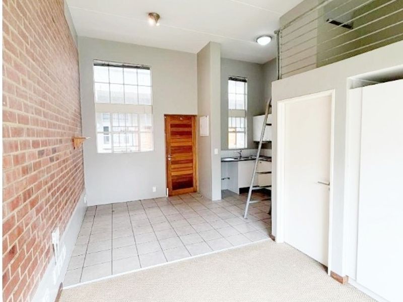 Open-plan living area featuring an exposed brick wall, mezzanine level with metal balustrade, and a compact kitchenette with built-in cabinetry.