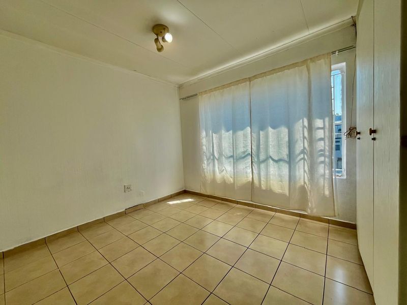 A bright, empty bedroom featuring tiled flooring, built-in cupboards, and security bars on the window.