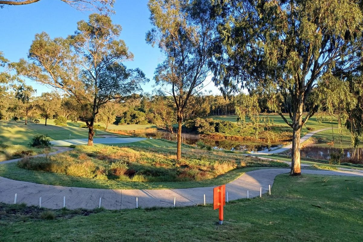 A scenic view overlooking a golf course with a water feature, mature trees, and a paved pathway winding through the landscape.