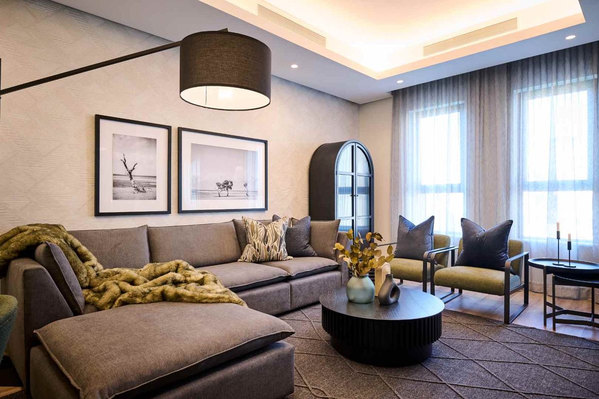 A modern, well-lit living room featuring a recessed tray ceiling with integrated lighting, large windows with sheer curtains, and textured wall finishes.