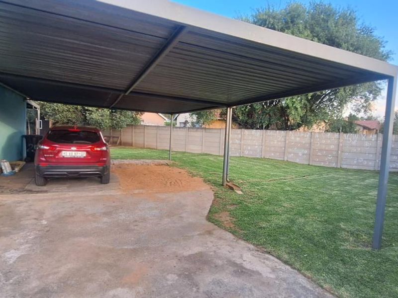 Rear exterior area featuring a large metal carport, concrete driveway, and a well-maintained lawn enclosed by precast concrete and metal fencing.