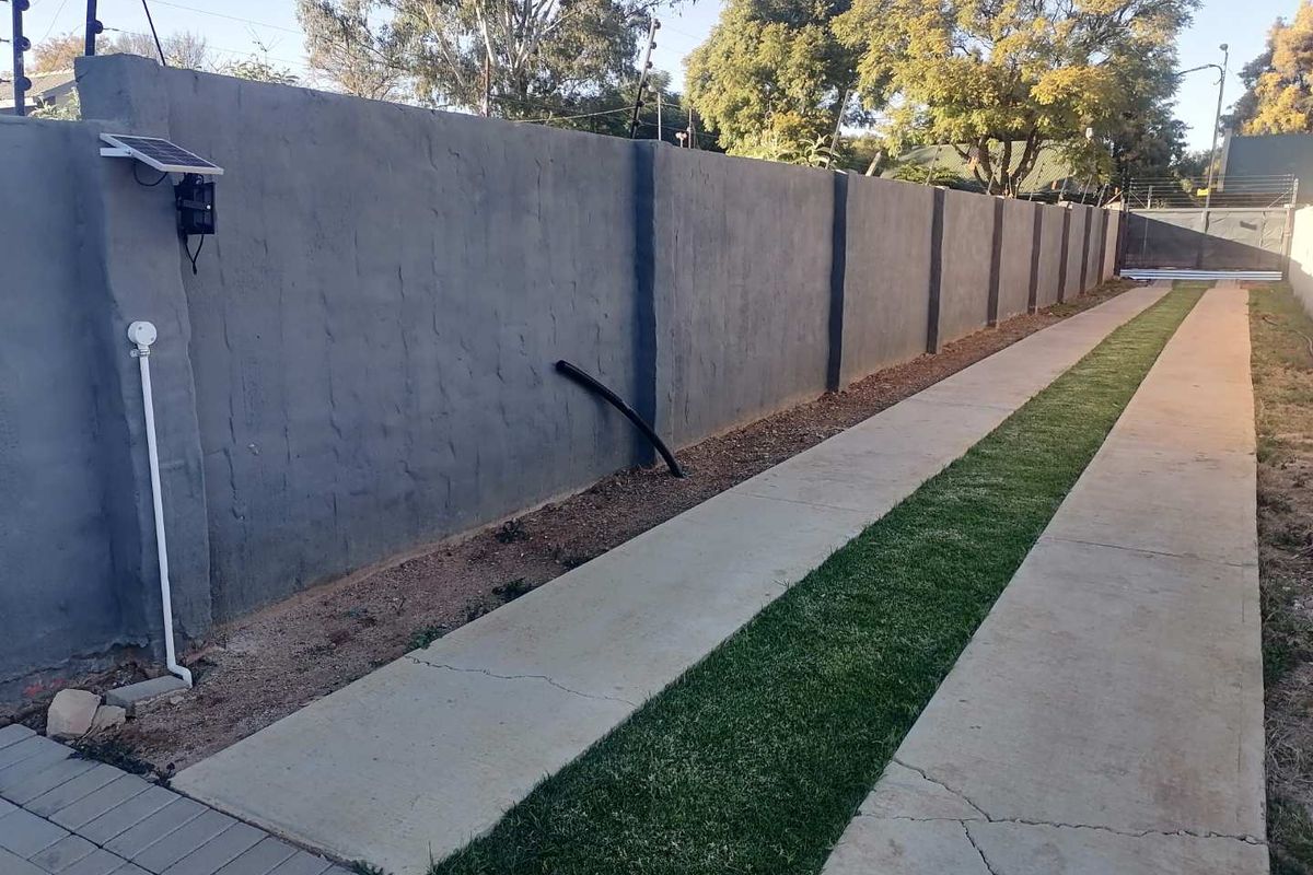 A side exterior view featuring a long concrete driveway with a central grass strip, bordered by a high perimeter boundary wall with electric fencing.