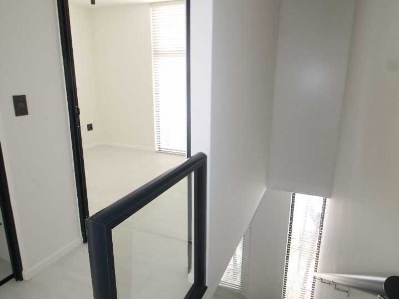 A modern, minimalist hallway featuring a staircase with a glass balustrade, clean white walls, and recessed lighting, leading to an adjacent room.