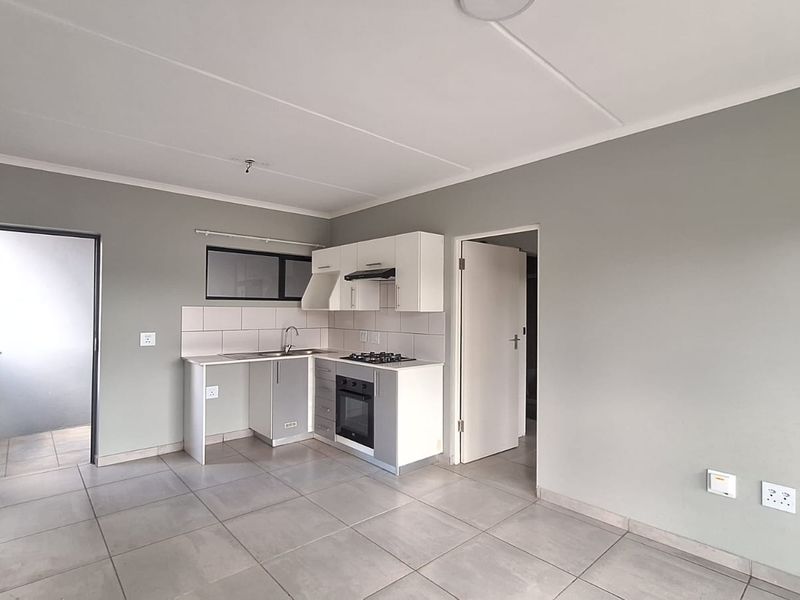 An open-plan living and kitchen area featuring neutral grey walls, tiled flooring, and a compact kitchen equipped with built-in cabinetry and appliances.