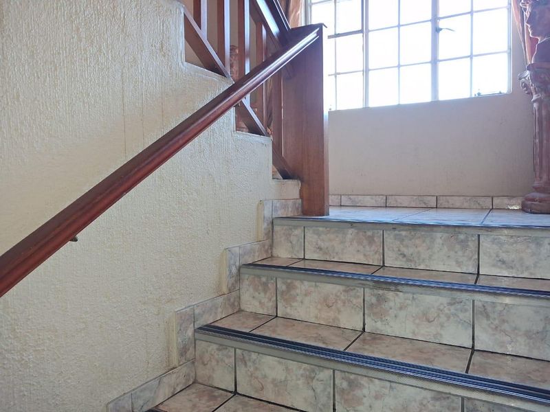 A staircase featuring tiled steps with safety nosing and a wooden handrail, illuminated by a large grid window.