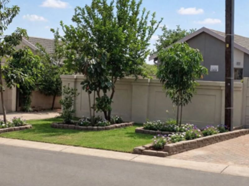 A well-maintained street-facing view featuring a neat boundary wall, landscaped garden beds with established trees, and a paved driveway entrance.