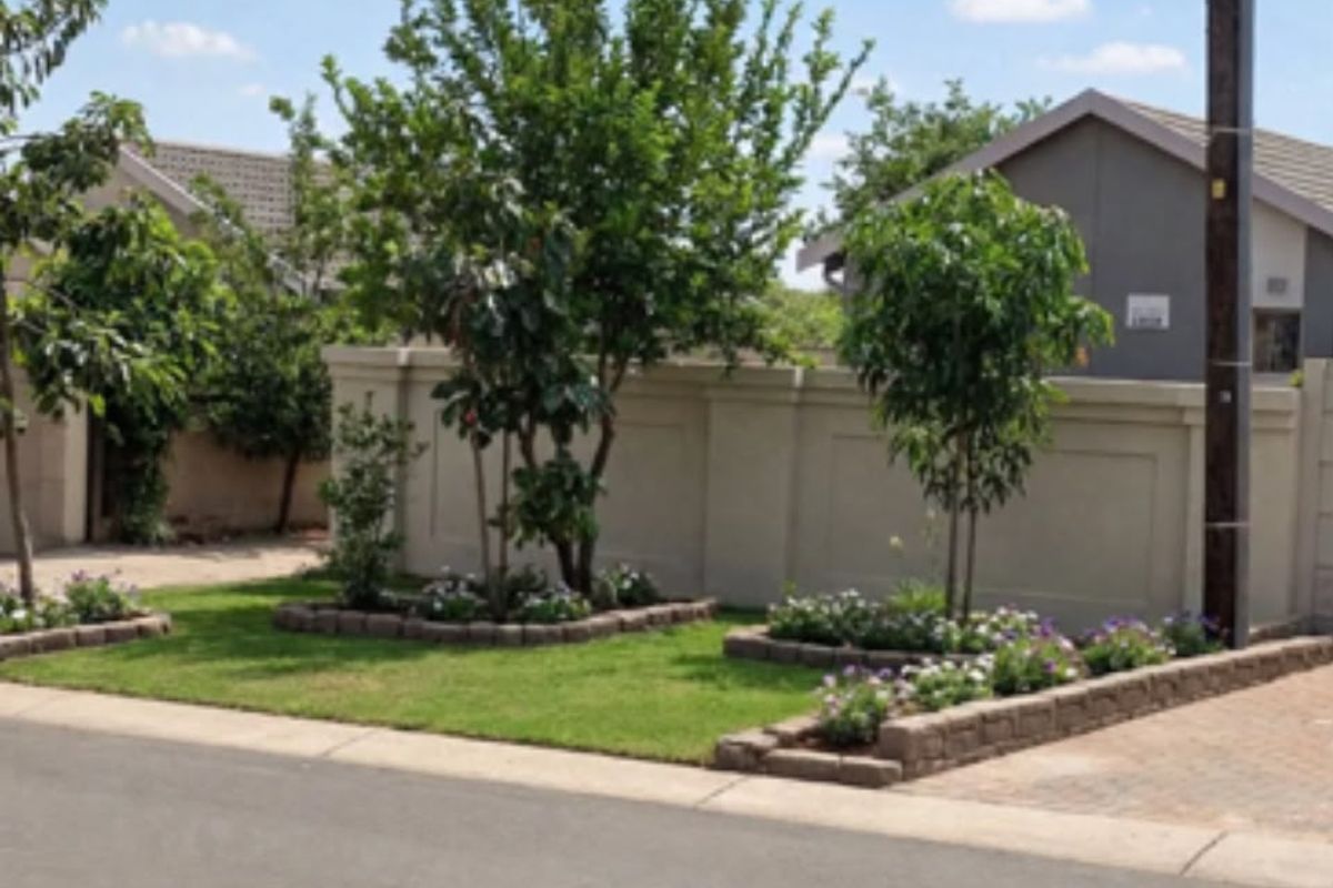 A well-maintained street-facing view featuring a neat boundary wall, landscaped garden beds with established trees, and a paved driveway entrance.