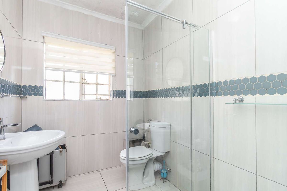 A functional bathroom featuring a pedestal basin, a glass-enclosed shower, and a toilet, finished with neutral wall tiles and a decorative mosaic border.