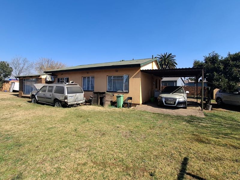 Rear view of the property featuring a single carport, concrete driveway, and a large garden area with established trees.