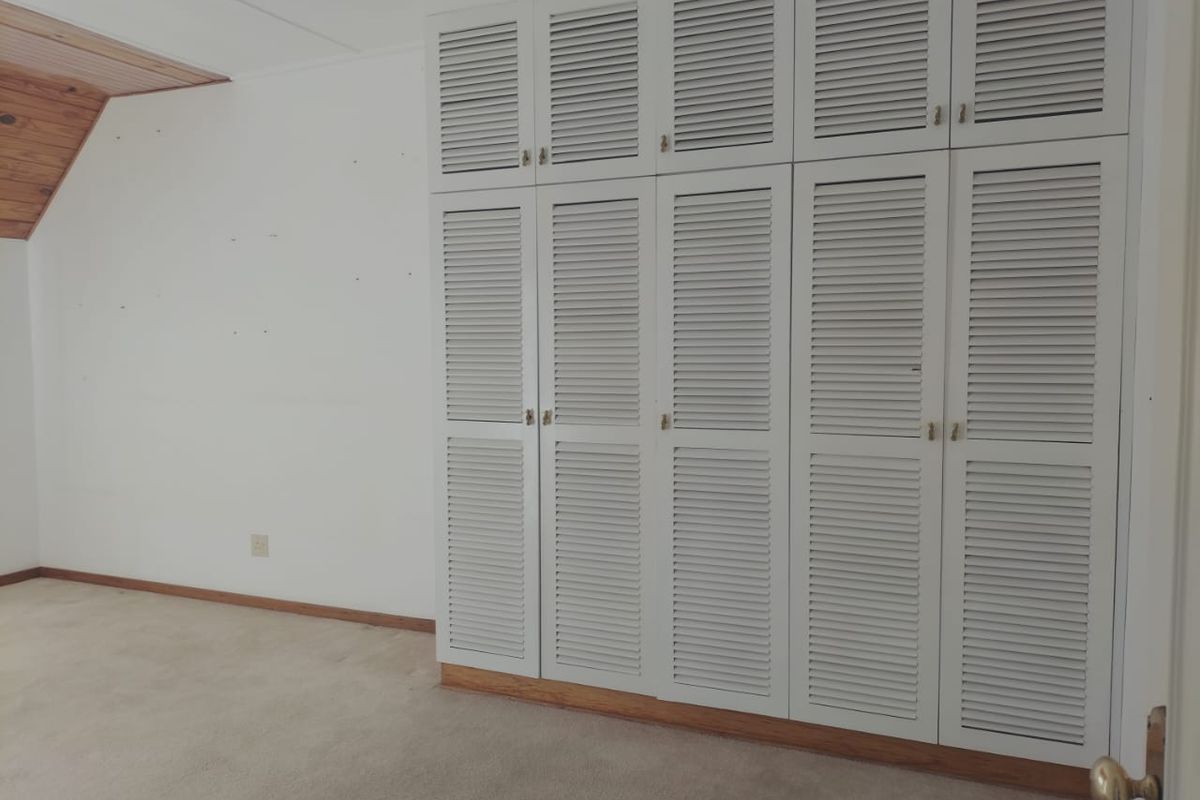 A bedroom featuring built-in white louvered cupboards, neutral carpeting, and a sloped timber-paneled ceiling section.