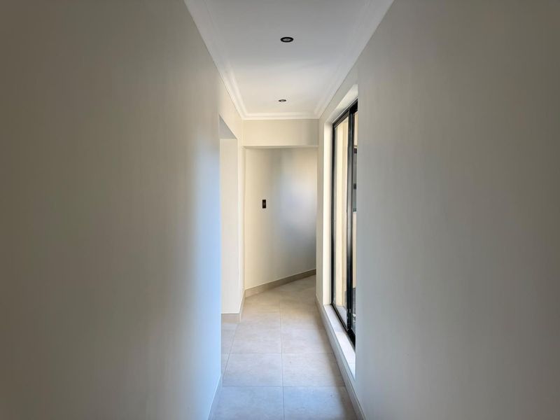 A bright, neutral-toned hallway featuring tiled flooring, recessed downlighting, and a large sliding glass door providing natural light.