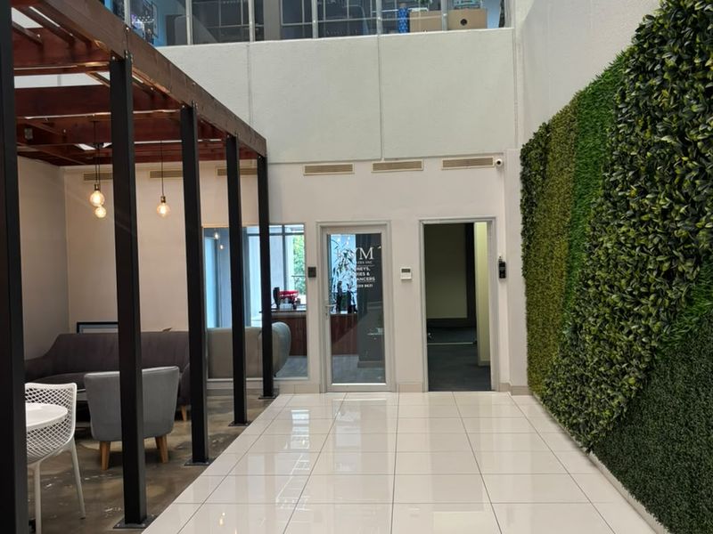A modern, double-volume open-plan office lobby featuring a vertical garden wall, timber pergola structure, and large glass windows providing ample natural light.