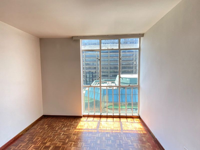 A bright, empty bedroom featuring classic parquet flooring and a large window with urban views, offering a blank canvas for personalization.