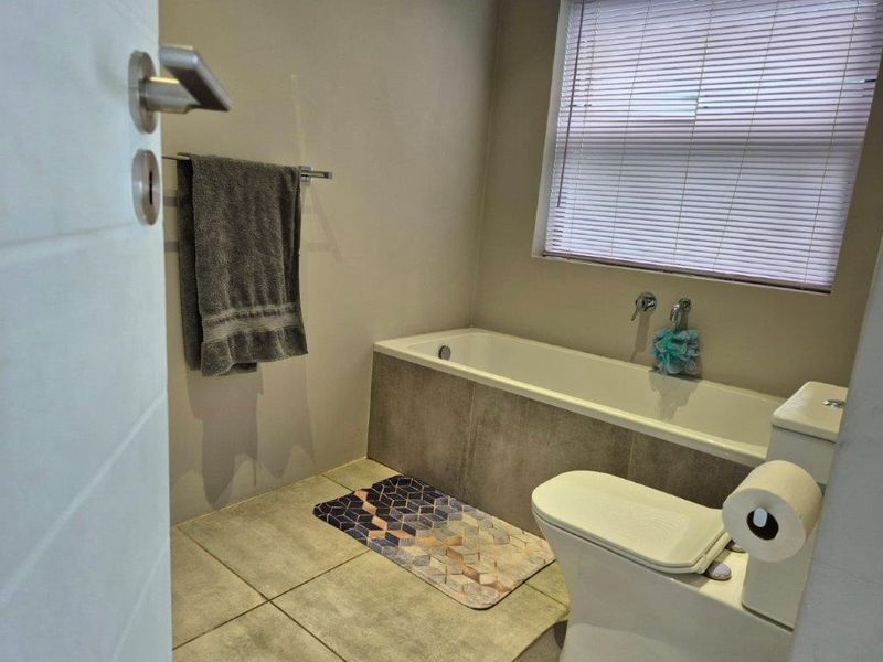 A functional bathroom featuring a built-in bathtub with tiled surround, a standard toilet, and neutral floor tiling with natural light from a window.