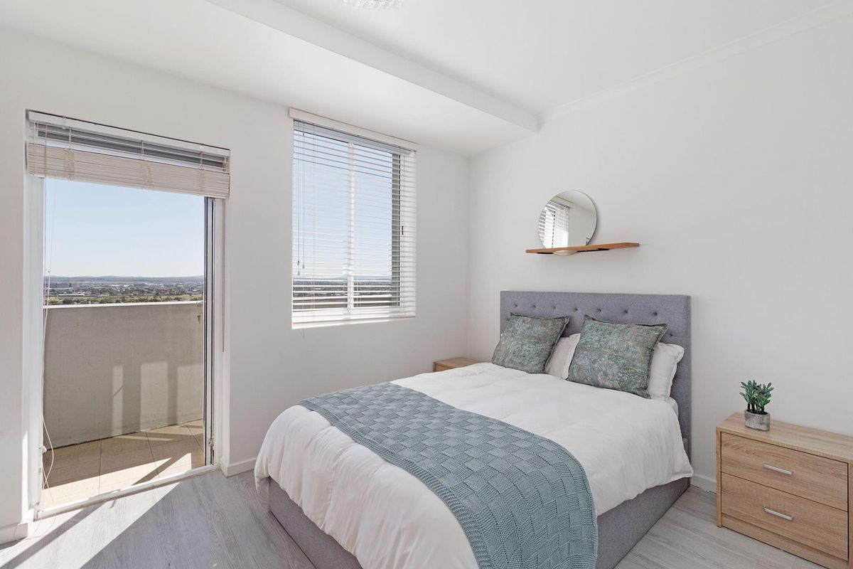 A bright and modern bedroom featuring light laminate flooring, venetian blinds, and a sliding glass door providing access to a private balcony.