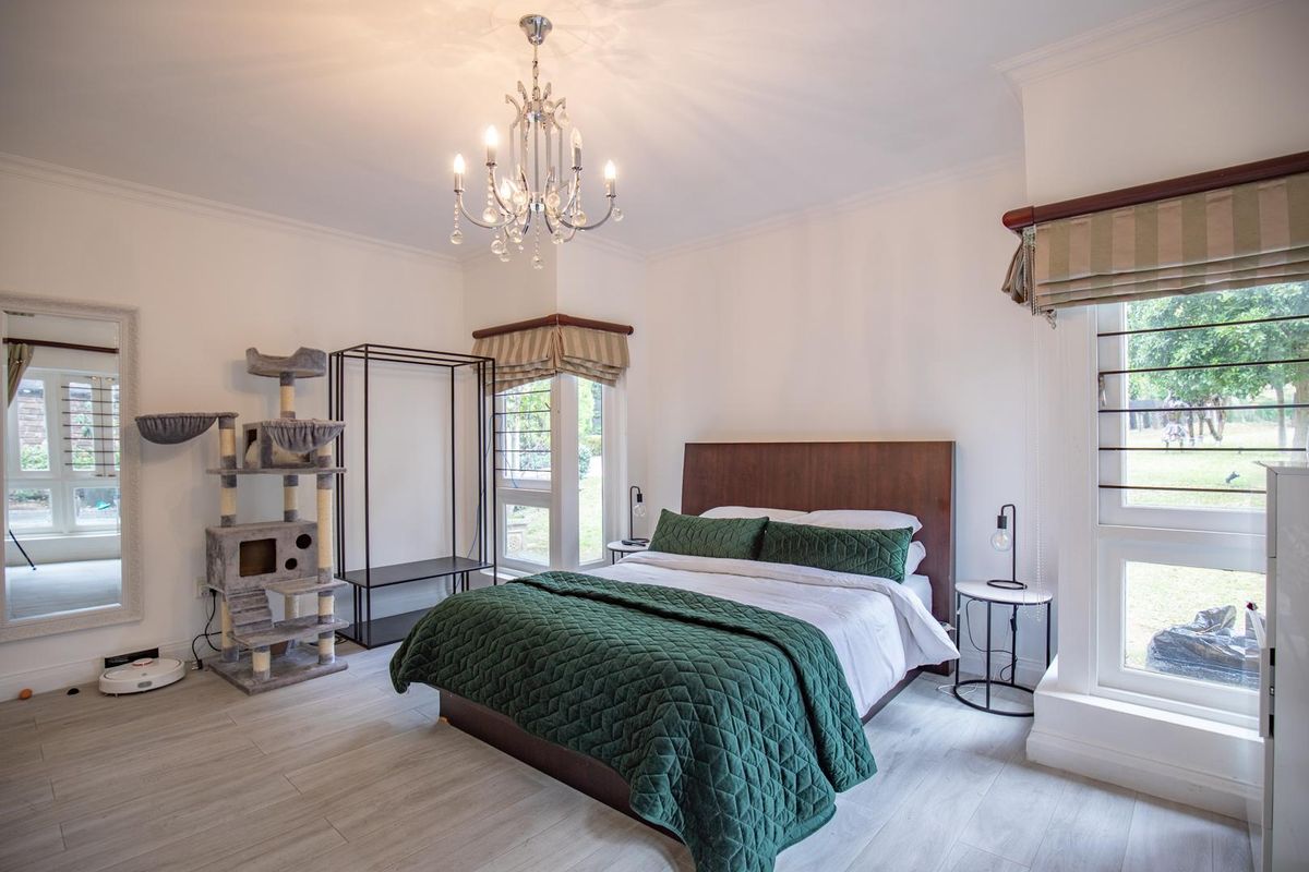 A bright bedroom featuring light-toned wood-look flooring, large windows with fitted blinds, and a decorative crystal chandelier.