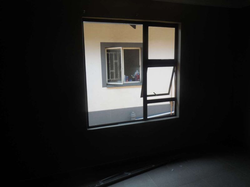 An empty, dimly lit room featuring tiled flooring and an aluminium window frame looking out onto an adjacent building exterior.