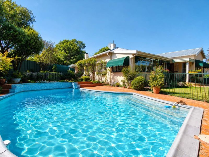 A well-maintained swimming pool area featuring a tiled pool, brick paved surround, and metal fencing, set against the rear elevation of the house.