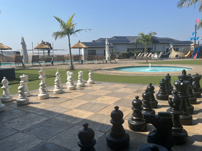 Outdoor recreational area featuring a large chess board, swimming pool, water fountain, and landscaped grounds with solar-equipped buildings in the background.