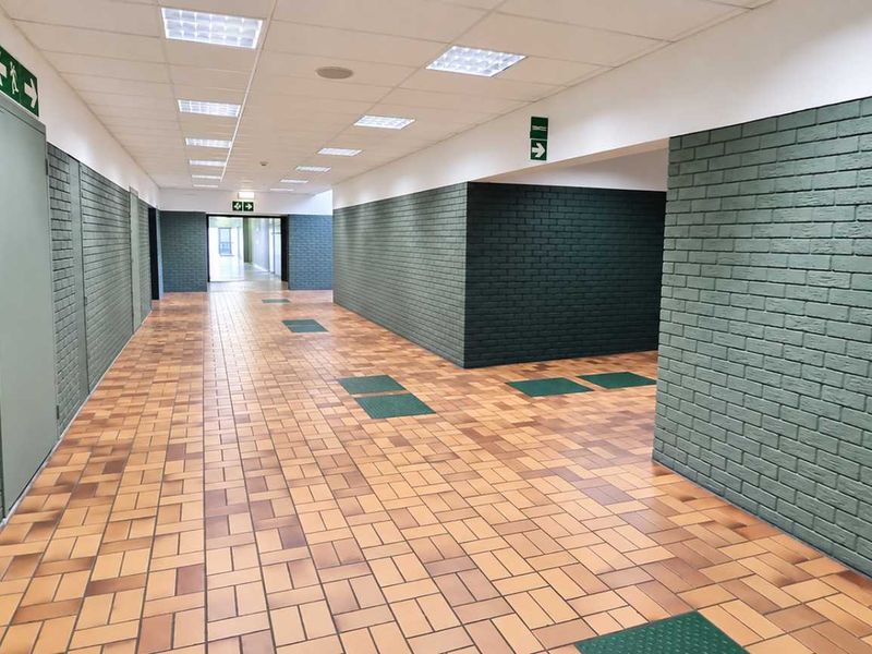 A spacious, well-maintained industrial hallway featuring durable ceramic tile flooring, painted brick walls, and integrated recessed ceiling lighting with clear emergency exit signage.