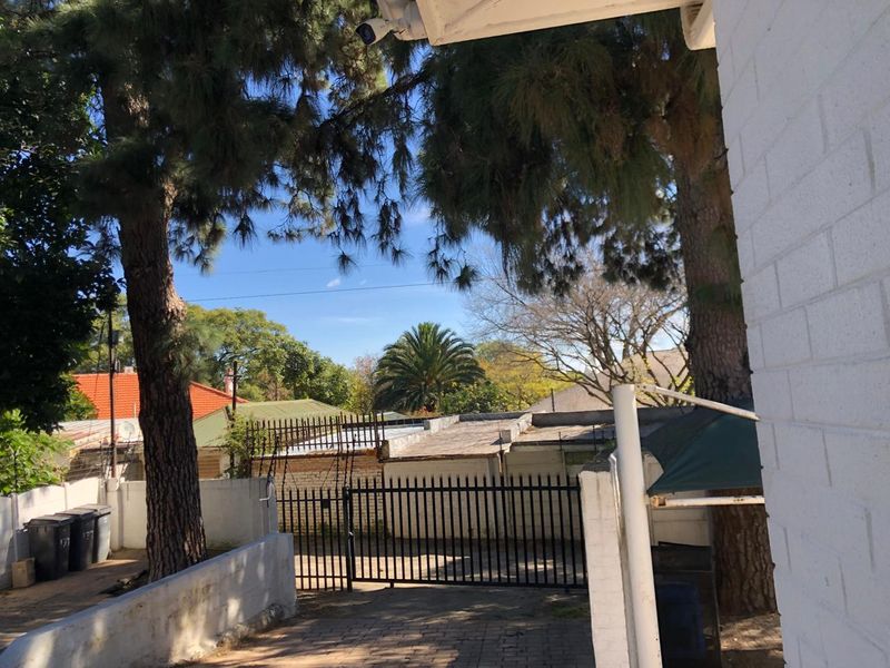 A view of the rear exterior area featuring a paved driveway, perimeter wall, and security gate, surrounded by mature trees.