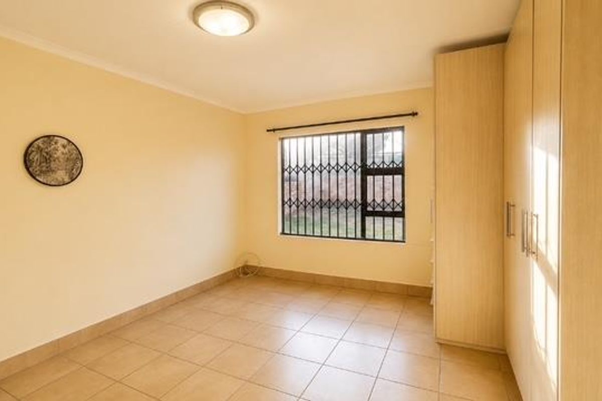 A bright, empty bedroom featuring neutral tiled flooring, built-in wooden cupboards, and a window fitted with security bars.