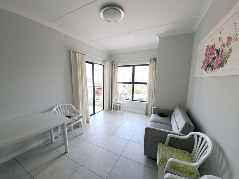 A bright living area featuring neutral tiled flooring, large windows, and a sliding door providing access to an outdoor balcony space.