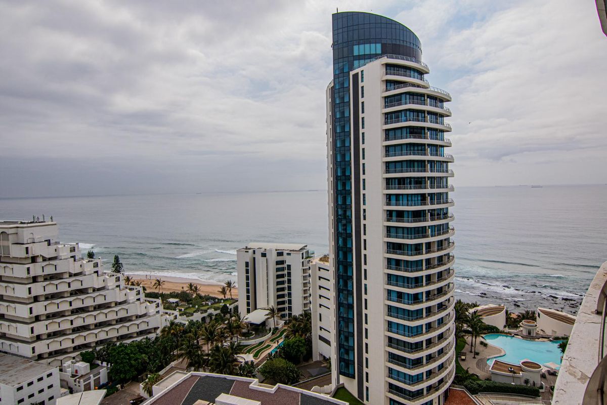 Aerial view of a modern high-rise apartment building situated along the coastline, featuring extensive balconies and proximity to the ocean.