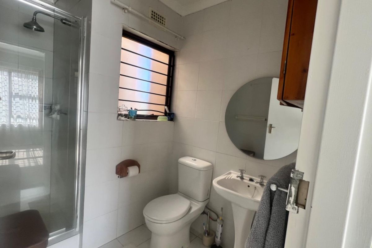 A functional bathroom featuring a shower enclosure, pedestal basin, and toilet with white wall tiling and security bars on the window.
