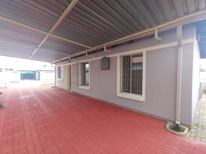 A covered outdoor patio area featuring paved flooring, a corrugated metal roof, and security-fitted windows along the exterior wall.