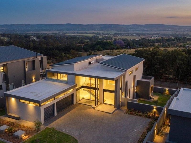 Aerial view of a modern contemporary residence featuring a double garage, paved driveway, and expansive glass facade at dusk.