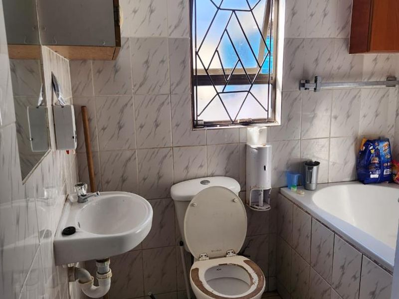 A functional bathroom featuring a bathtub, pedestal basin, and toilet, finished with ceramic wall and floor tiles and fitted with security burglar bars.