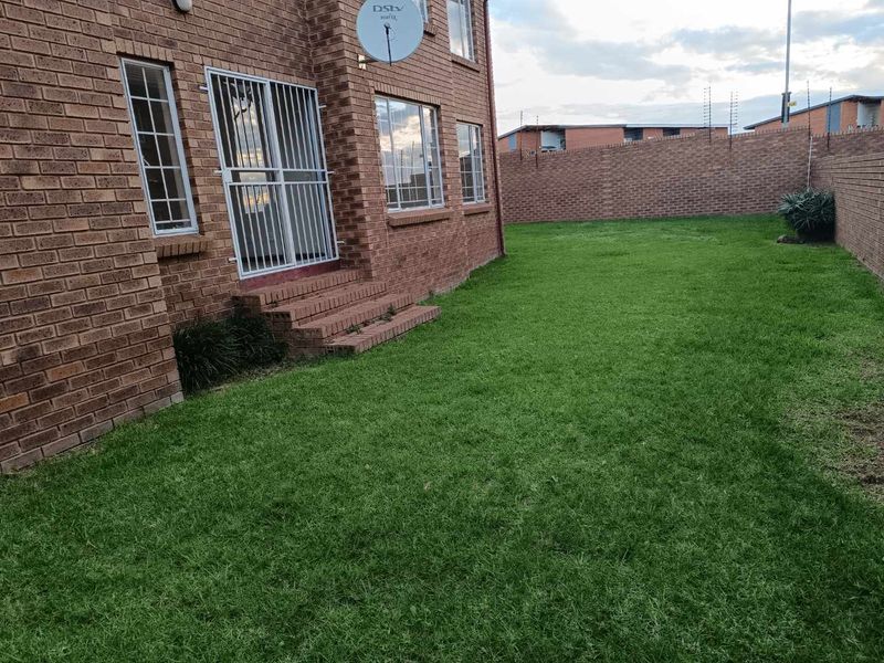 Rear view of the townhouse featuring a face brick exterior, a small lawn area, and a secure entrance with a metal security gate.