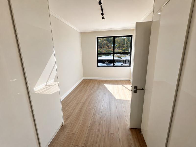 A bright, empty bedroom featuring modern laminate flooring, built-in cupboards with high-gloss finish, and a large window with black frames.