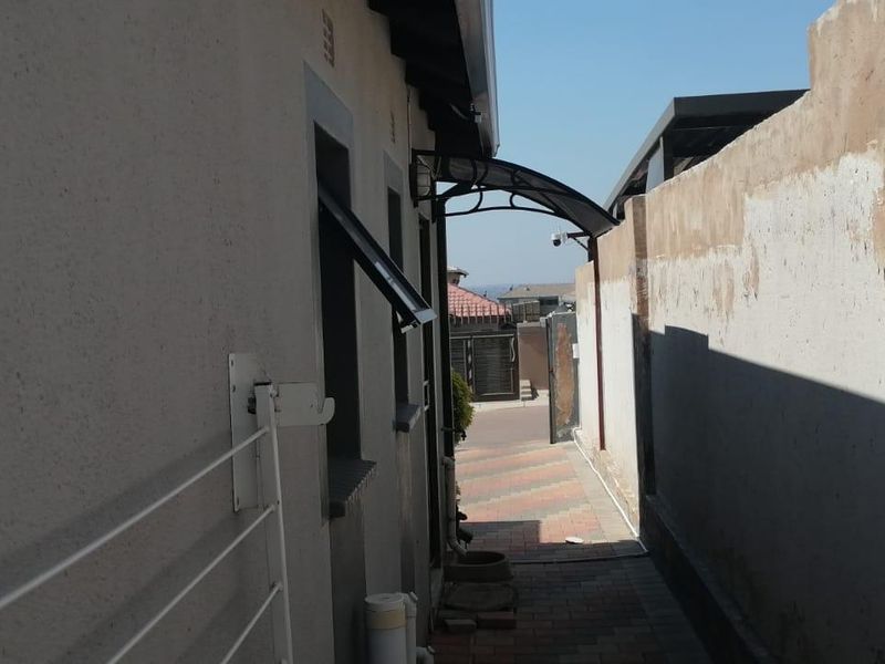 A narrow side passage featuring a paved walkway, a protective awning, and a boundary wall requiring maintenance.