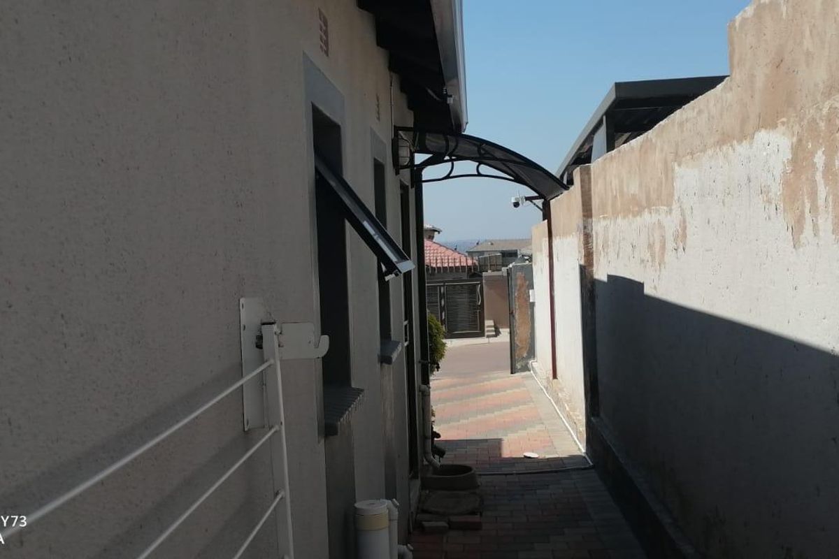 A narrow side passage featuring a paved walkway, a protective awning, and a boundary wall requiring maintenance.