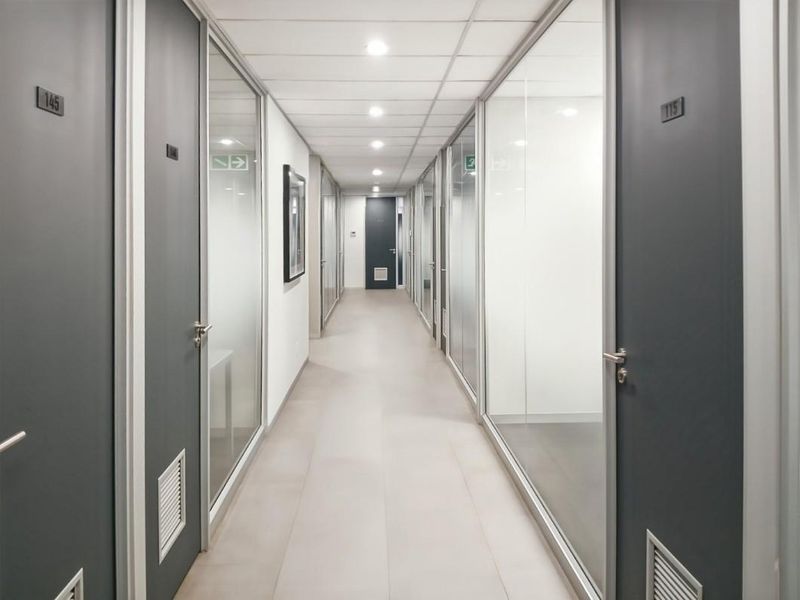 A modern office hallway featuring glass partition walls, tiled flooring, and recessed ceiling lighting, providing a clean and professional corridor access to individual office spaces.